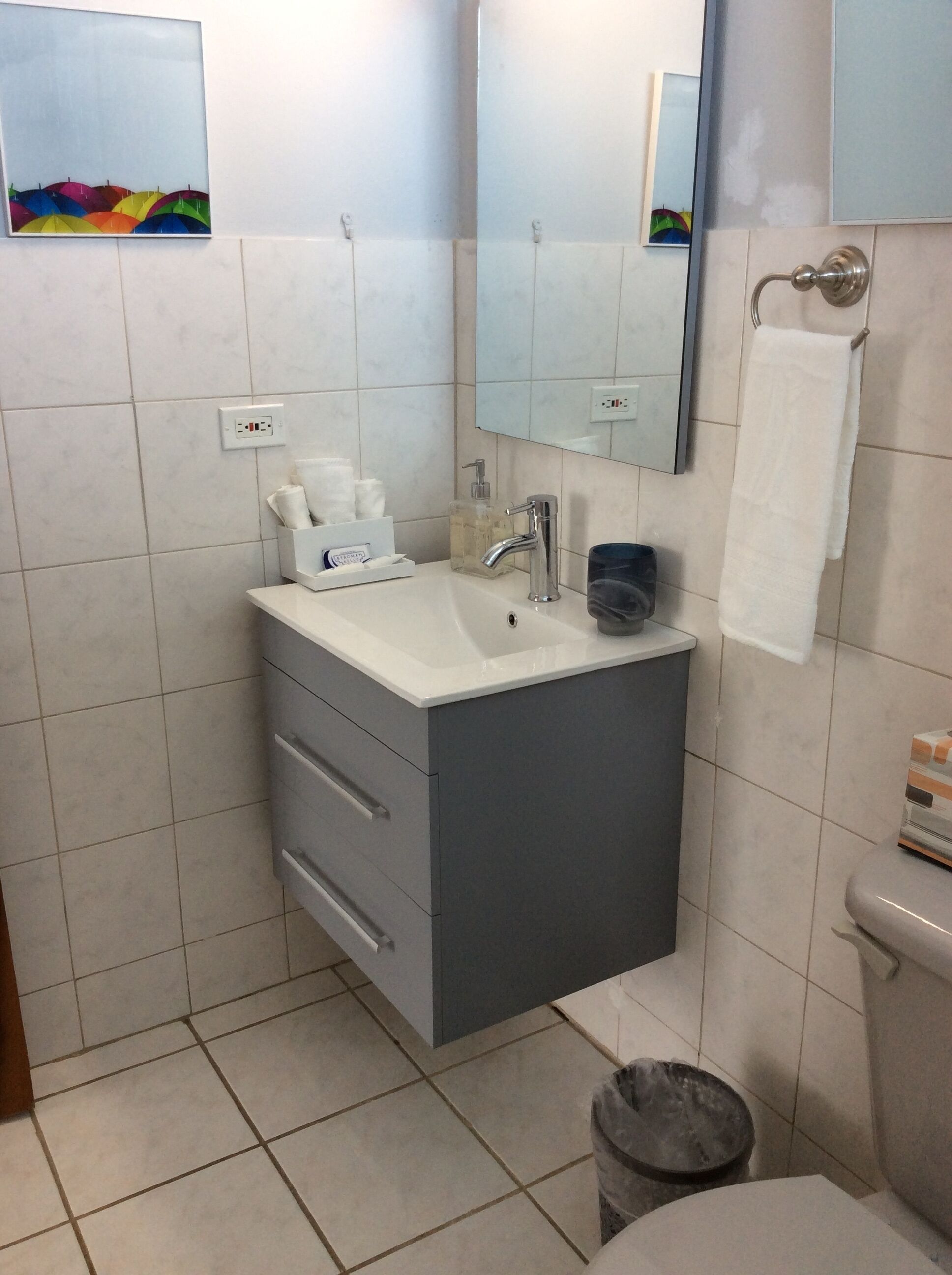 Een badkamer met een witte wastafel, een spiegel, een handdoekrek en een prullenbak.