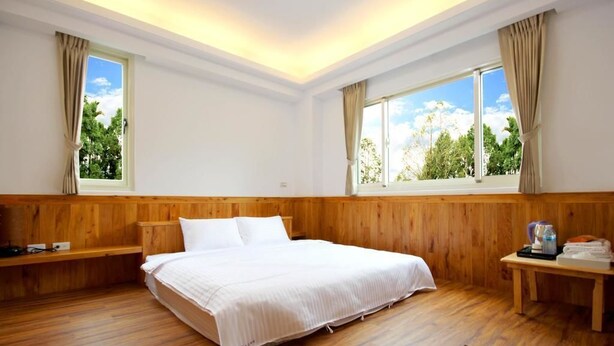 Quarto de hotel com cama de madeira, duas janelas com cortinas, mesa de cabeceira com chaleira e vista para o verde lá fora.
