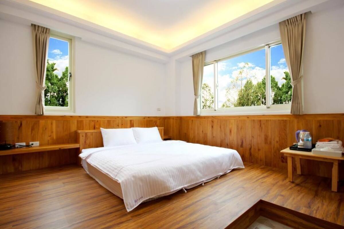 Ein Hotelzimmer mit einem hölzernen Bett, zwei Fenstern mit Vorhängen, einem Nachttisch mit Wasserkocher und Blick auf Grünflächen.