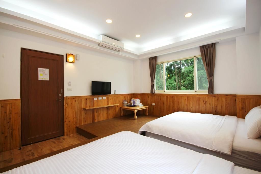 A hotel room with two beds, a television, a window with curtains, and a small table with a cup and a book.