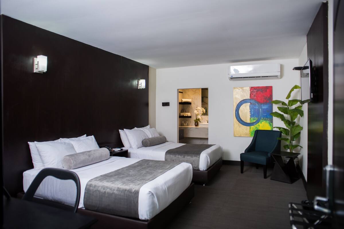 A hotel room with two beds, a desk, a chair, an air conditioning unit, and a colorful abstract painting.