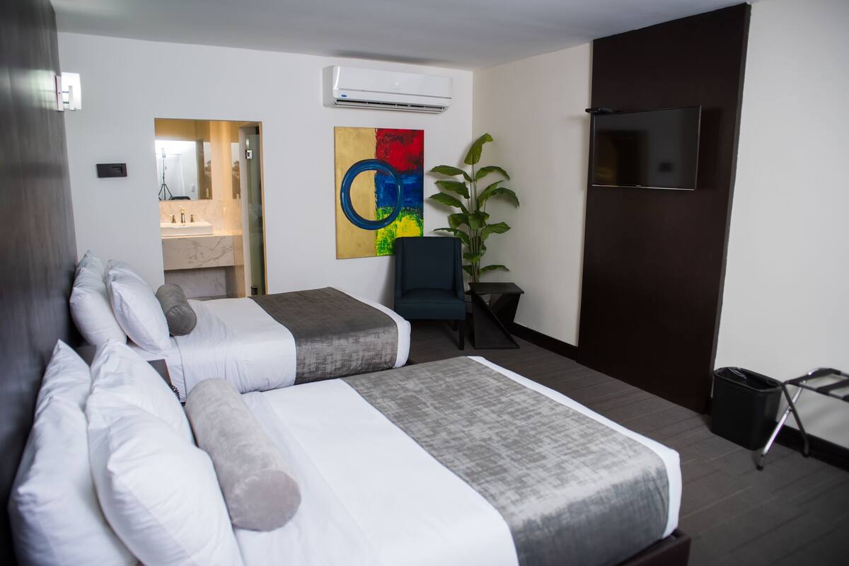 A hotel room with two beds, a TV, a plant, and a painting on the wall.