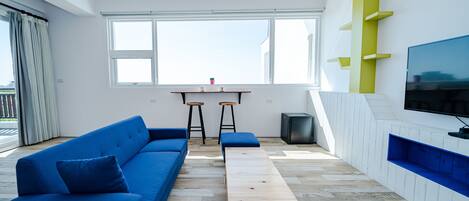 Uma sala de estar moderna com um sofá azul, uma mesa de centro de madeira e uma TV fixada em uma parede branca.