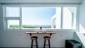 A room with a large window overlooking a beach, two stools, a small table, and a black appliance.