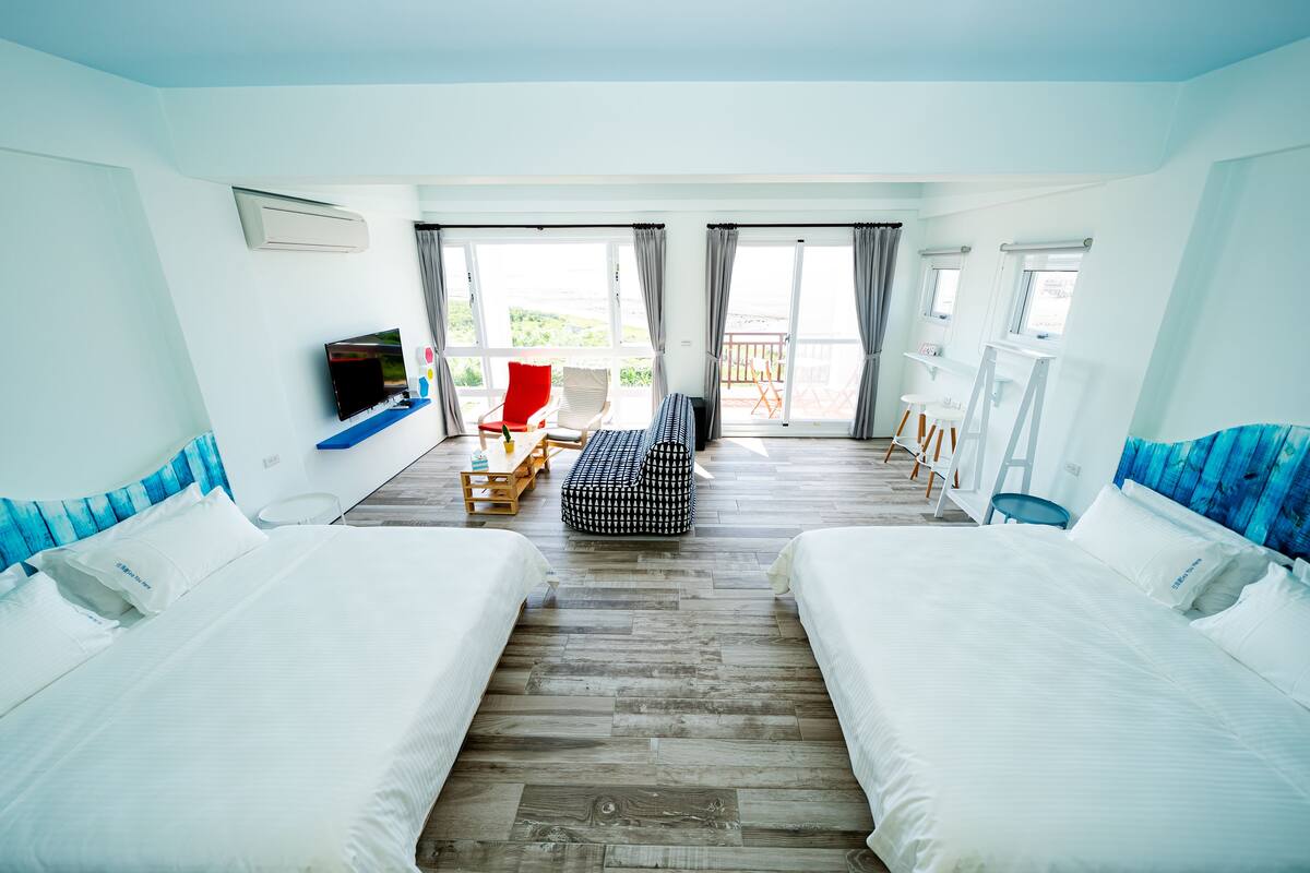 Hotellihuone, jossa on kaksi sänkyä, televisio, punainen tuoli, pieni pöytä ja parveke, jolta on näkymä.