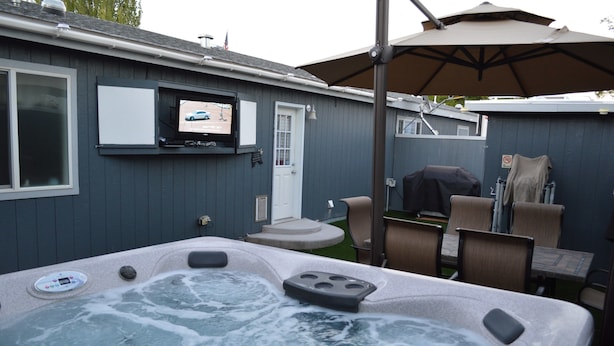 Un jardin avec un jacuzzi, un coin salon extérieur, un téléviseur fixé au mur et un espace barbecue couvert.
