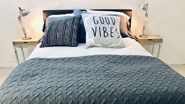 Un lit bien fait, recouvert d’une couette grise, avec deux coussins à motifs et un coussin orné de l’inscription « BONNE AMBIANCE ». Deux lampes de chevet sont posées sur des tables de nuit en bois.