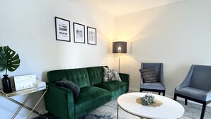 Un salon avec un canapé vert, deux fauteuils, une table basse, une lampe et des tableaux encadrés accrochés au mur.