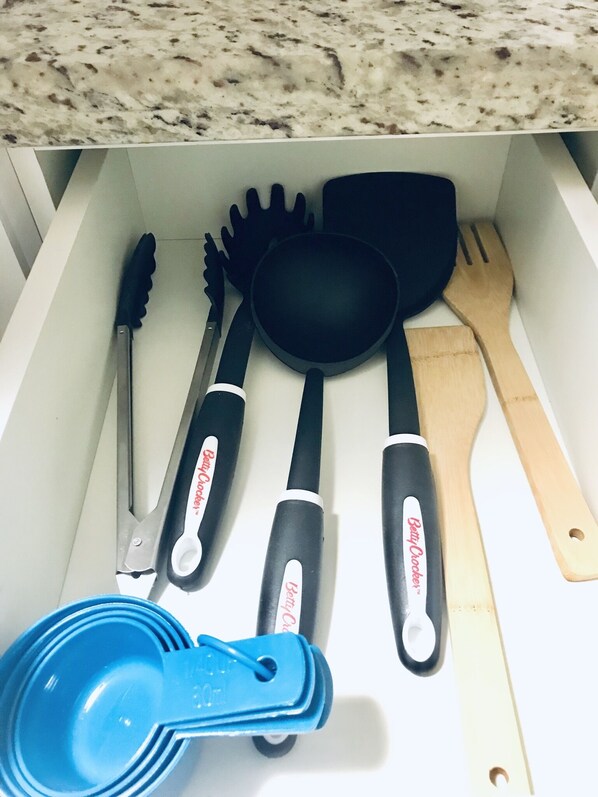 A drawer containing kitchen utensils including a spatula, a spoon, a ladle, a pair of tongs, and a measuring cup.