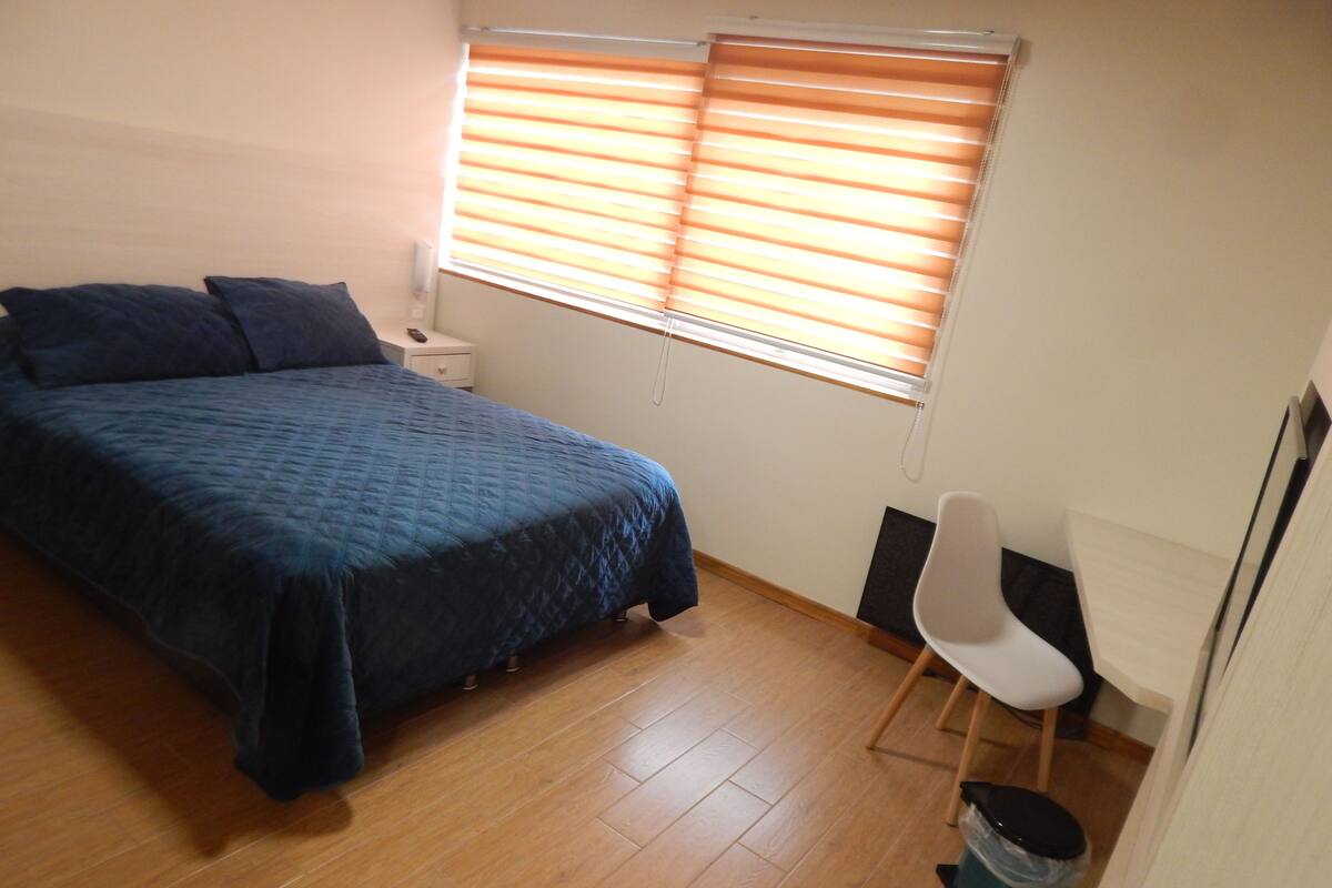 Ein Schlafzimmer mit Bett, Nachttisch, Stuhl, Fenster mit Jalousien und einem Mülleimer.