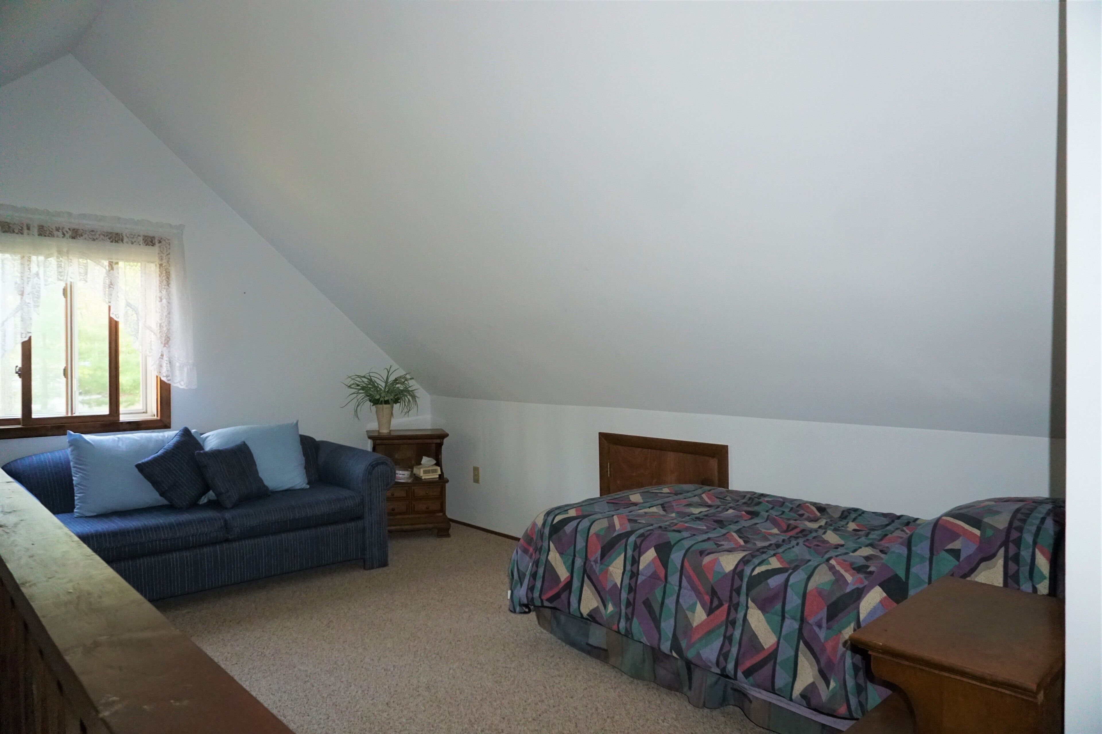 A bedroom with a sloped ceiling, a sofa, a bed with a colorful geometric-patterned bedspread, and a small wooden nightstand.