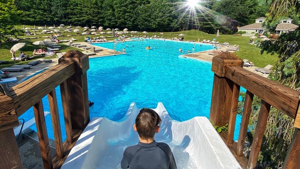Ein Kind saust eine Holzrutsche in ein großes Freibad, umgeben von Liegestühlen und Sonnenschirmen.