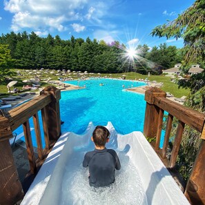 A child sliding down a wooden slide into a large outdoor swimming pool surrounded by lounge chairs and umbrellas.