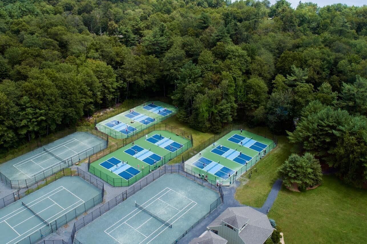 An aerial view of a tennis facility with multiple courts surrounded by dense forest.