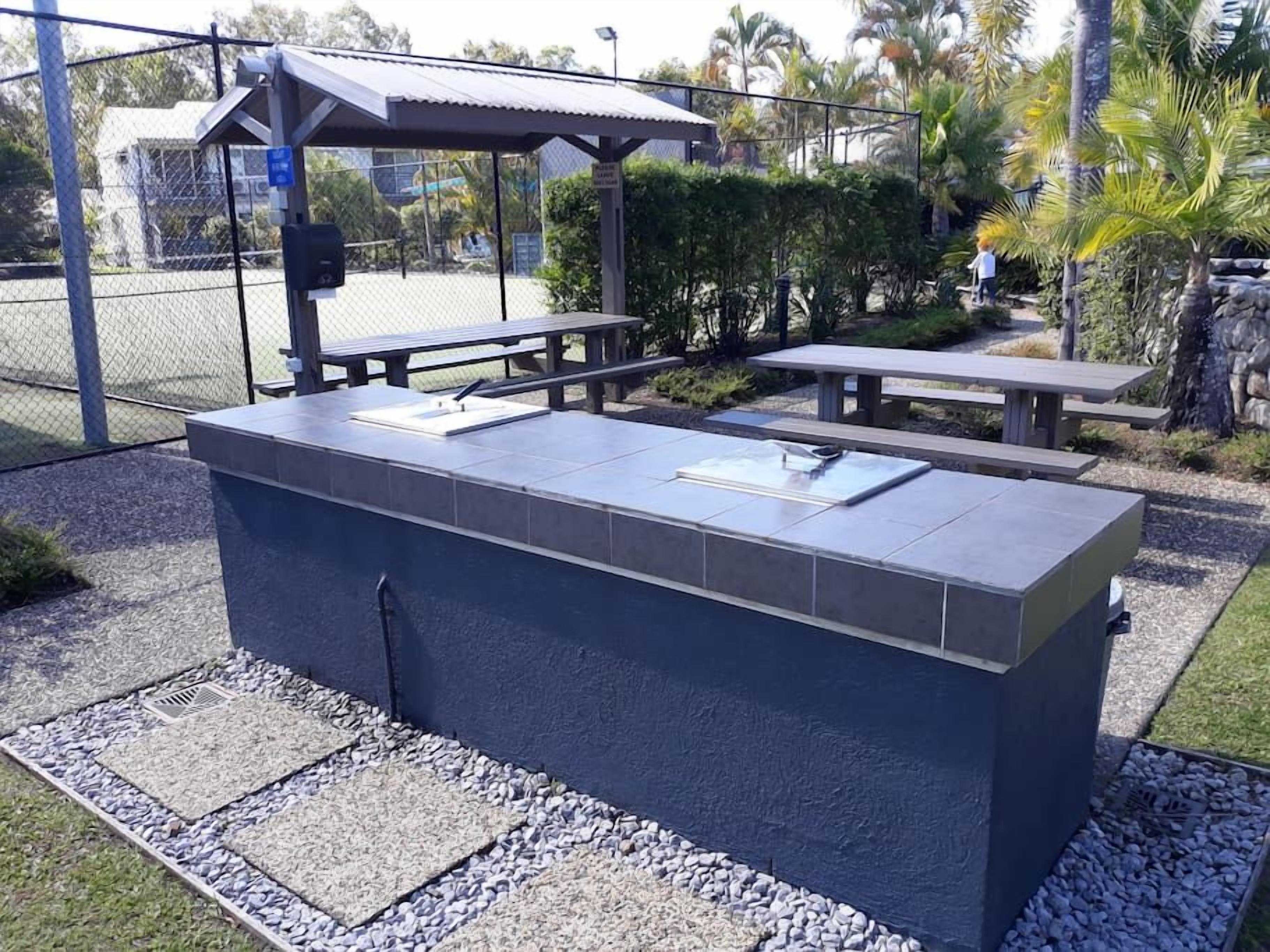 An outdoor seating area with a built-in table and benches, surrounded by a fence and palm trees.