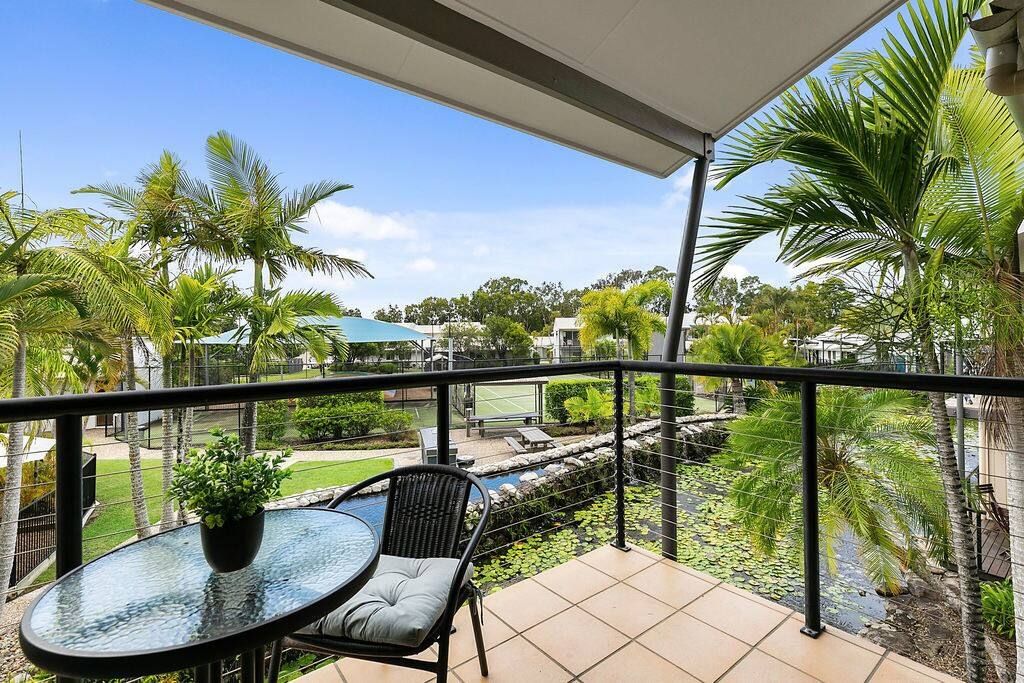 A balcony with a glass table and chair, overlooking a garden with palm trees and a small pond.