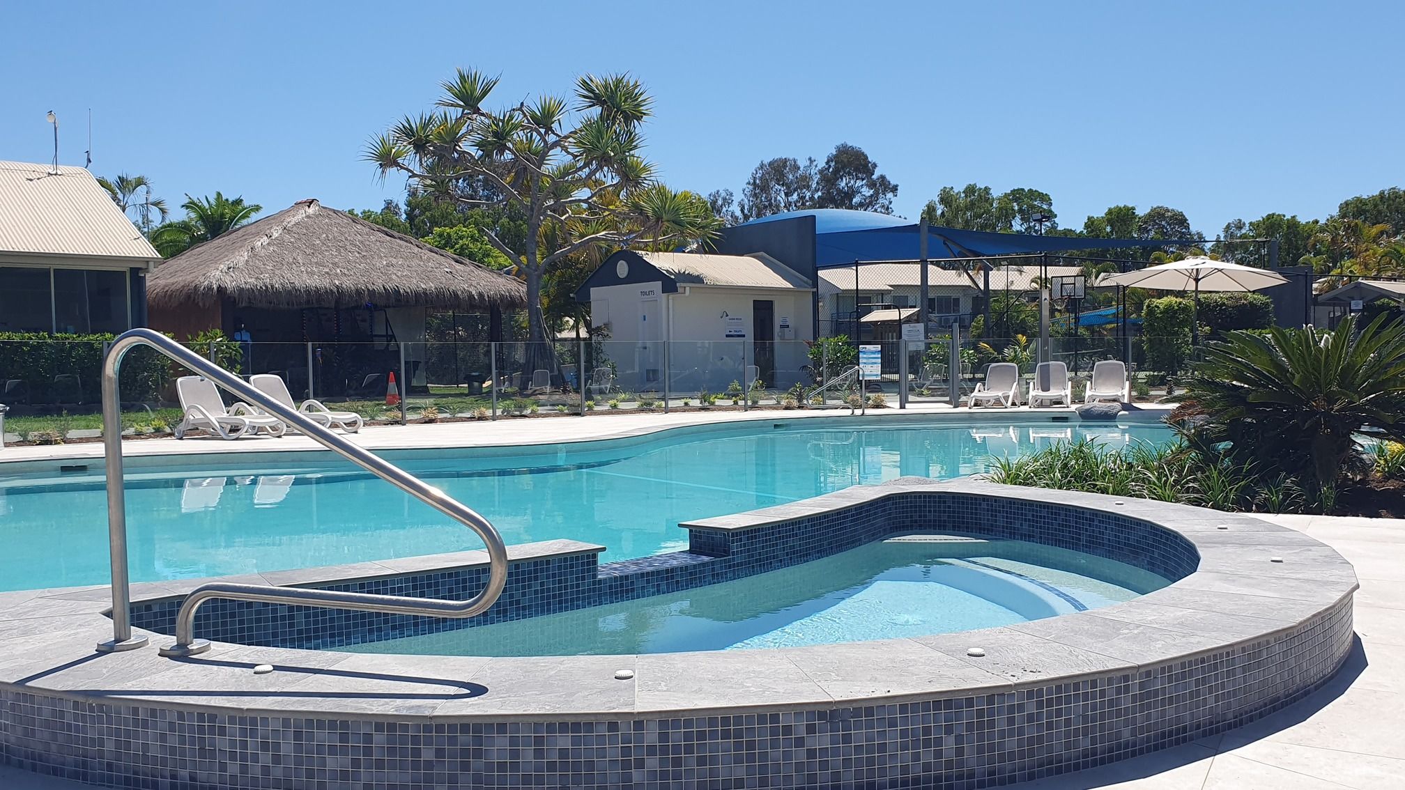 A swimming pool with a hot tub, lounge chairs, and a thatched-roof building.