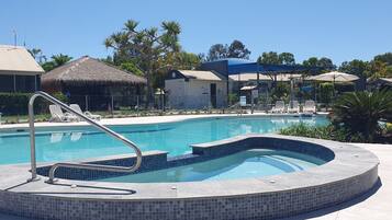 A swimming pool with a hot tub, lounge chairs, and a thatched-roof building.