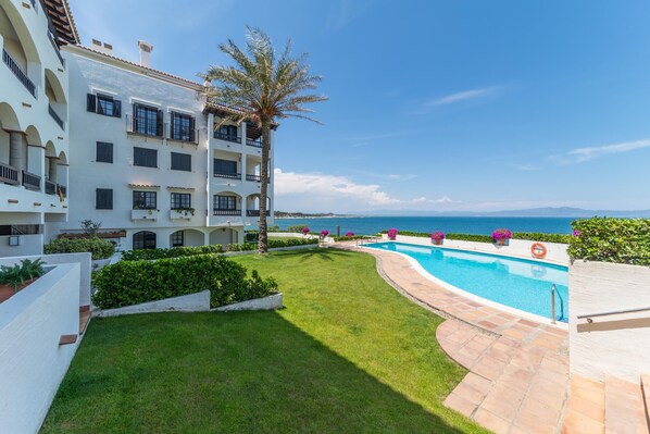 A poolside view with a clear blue sky, a white building with balconies, and a view of the sea.