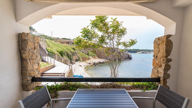 Un balcón con mesa y sillas, con vista a una zona costera con costa rocosa y un árbol.