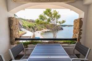 A balcony with a table and chairs, overlooking a coastal area with a rocky shoreline and a tree.