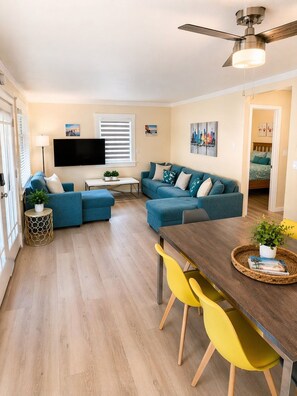 Un salon comprenant un ensemble de canapés, un téléviseur, un ventilateur de plafond et un coin repas avec des chaises jaunes.