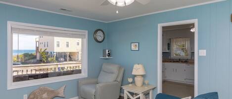 A living room with a view of the ocean, a ceiling fan, a clock, and a kitchen visible through an open doorway.