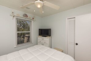 Un dormitorio con cama, televisor sobre una cómoda, ventilador de techo y ventana con persianas.