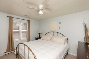 Quarto com cama, ventilador de teto, janela com persianas, mesa de cabeceira com abajur e um adorno na parede.