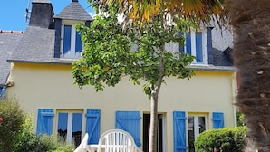 A two-story house with a yellow facade, blue shutters, and a white plastic chair in the yard.