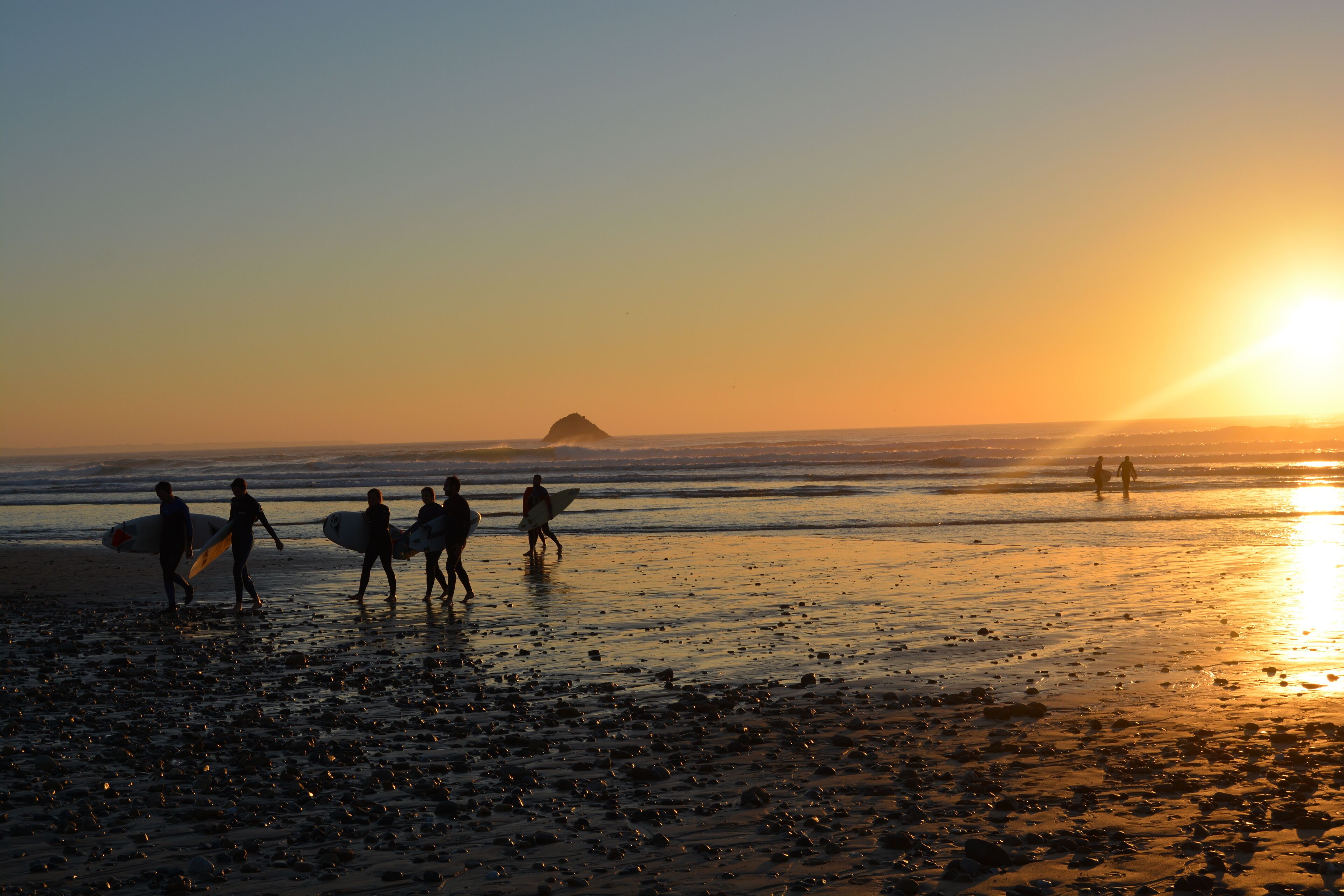 A beach scene with people walking and carrying surfboards, with the sun setting in the background.