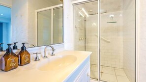 Ein Badezimmer mit einem weißen Waschbecken, zwei Shampoo-Flaschen, einem Spiegel und einer Duschkabine.