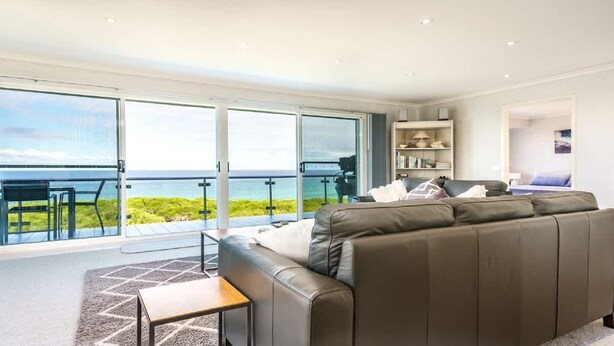 Una sala de estar moderna con un sofá grande, una mesita y una estantería. Hay un balcón con vistas al mar.