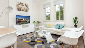 Ett modernt vardagsrum med en platt-TV, vita möbler, ett soffbord i glas och utsikt över grönområden utanför.