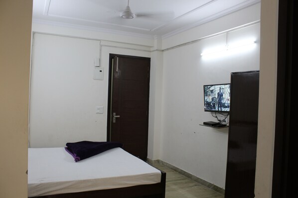 A small, sparsely furnished room with a bed, a door, a television on a stand, and a light fixture on the wall. - Hotel South Delhi Inn (New Delhi)
