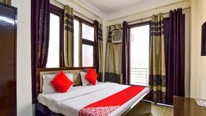 A hotel room with a bed, two red pillows, a red and white patterned bedspread, and curtains with a geometric design. - Hotel South Delhi Inn (New Delhi)