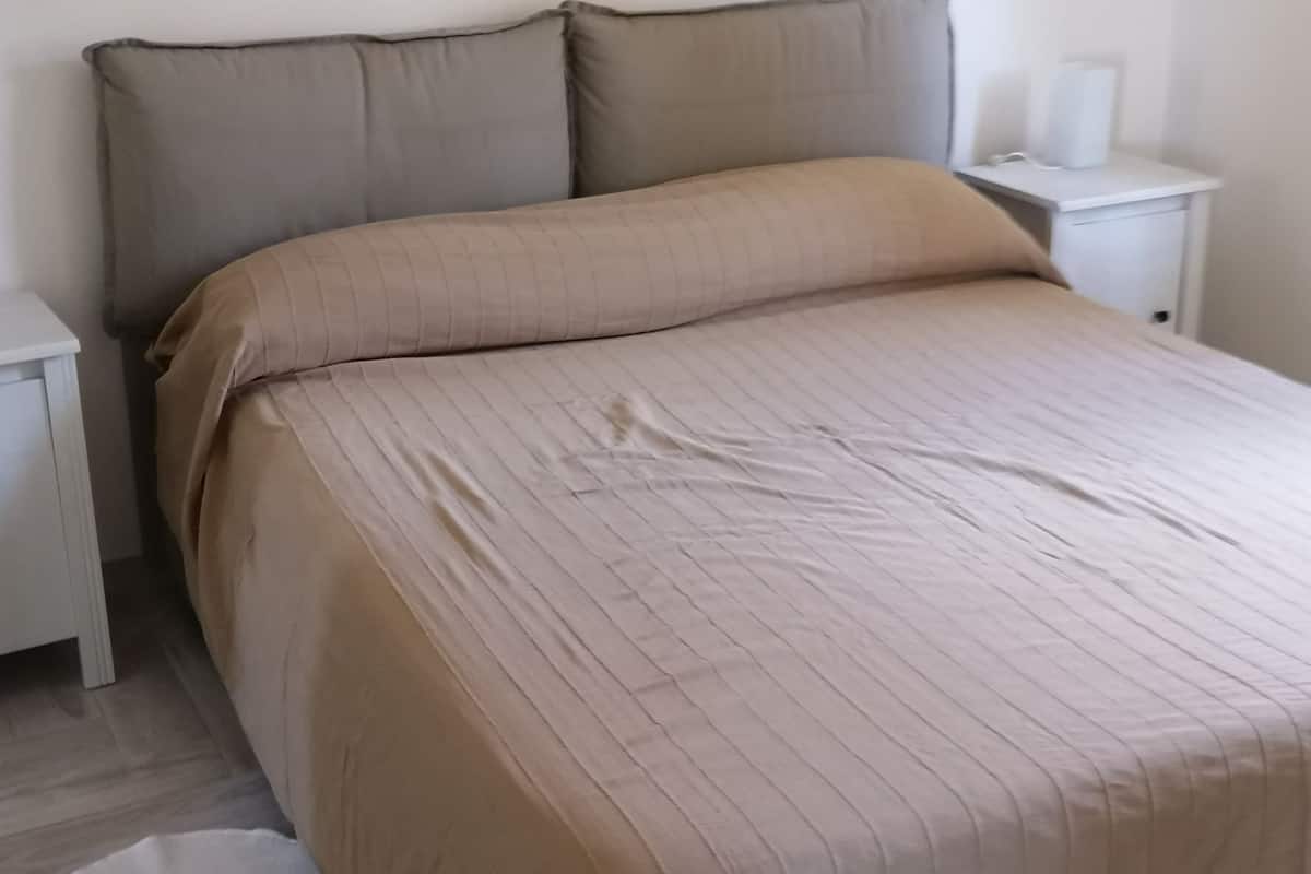 A neatly made bed with a beige bedspread and pillows, a small bedside table with a lamp, and a framed picture on the wall.