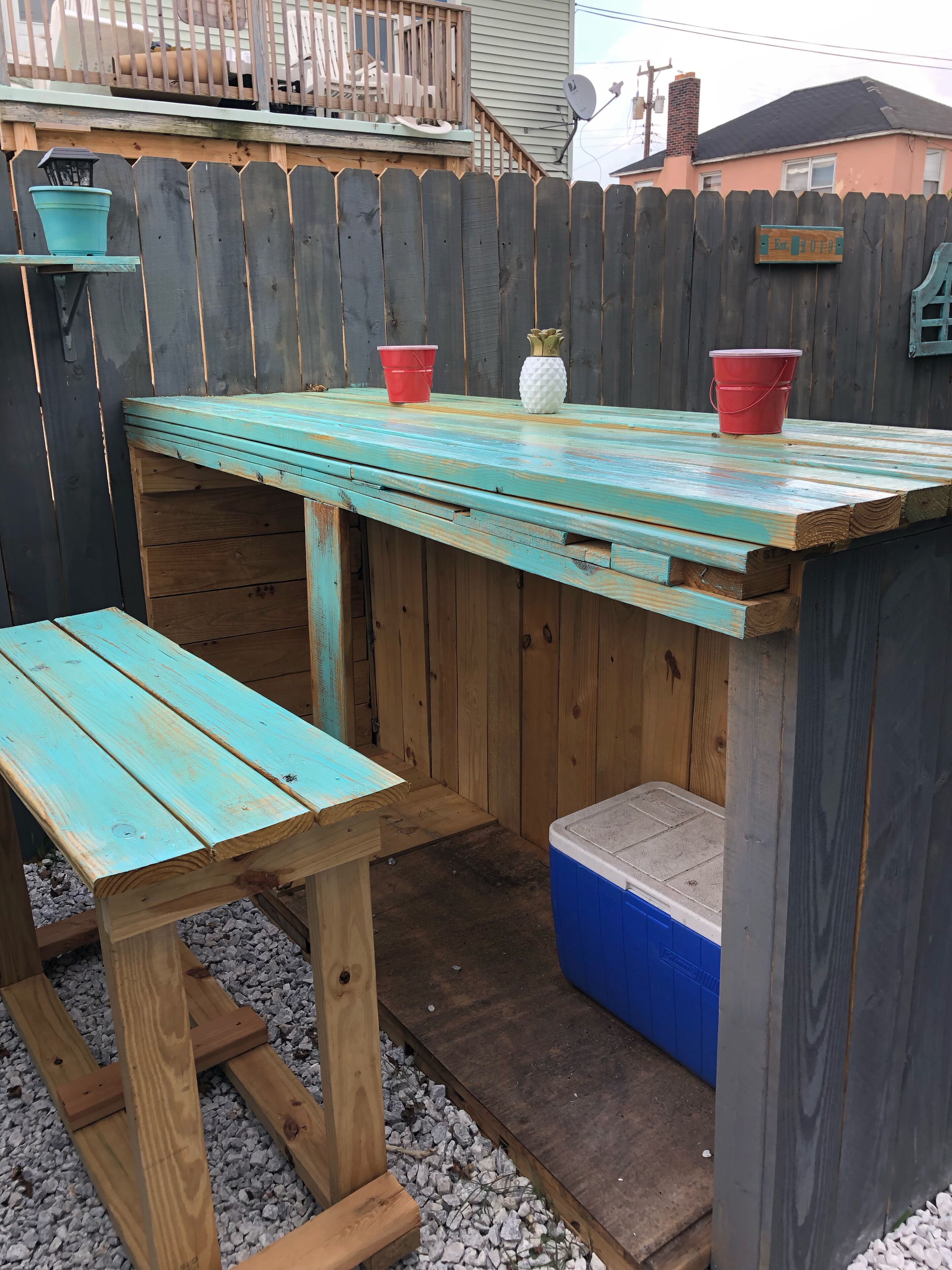 A wooden outdoor seating area with a table and benches, a small cooler, and a wooden fence.