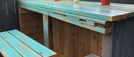 A wooden outdoor seating area with a table and benches, a small cooler, and a wooden fence.