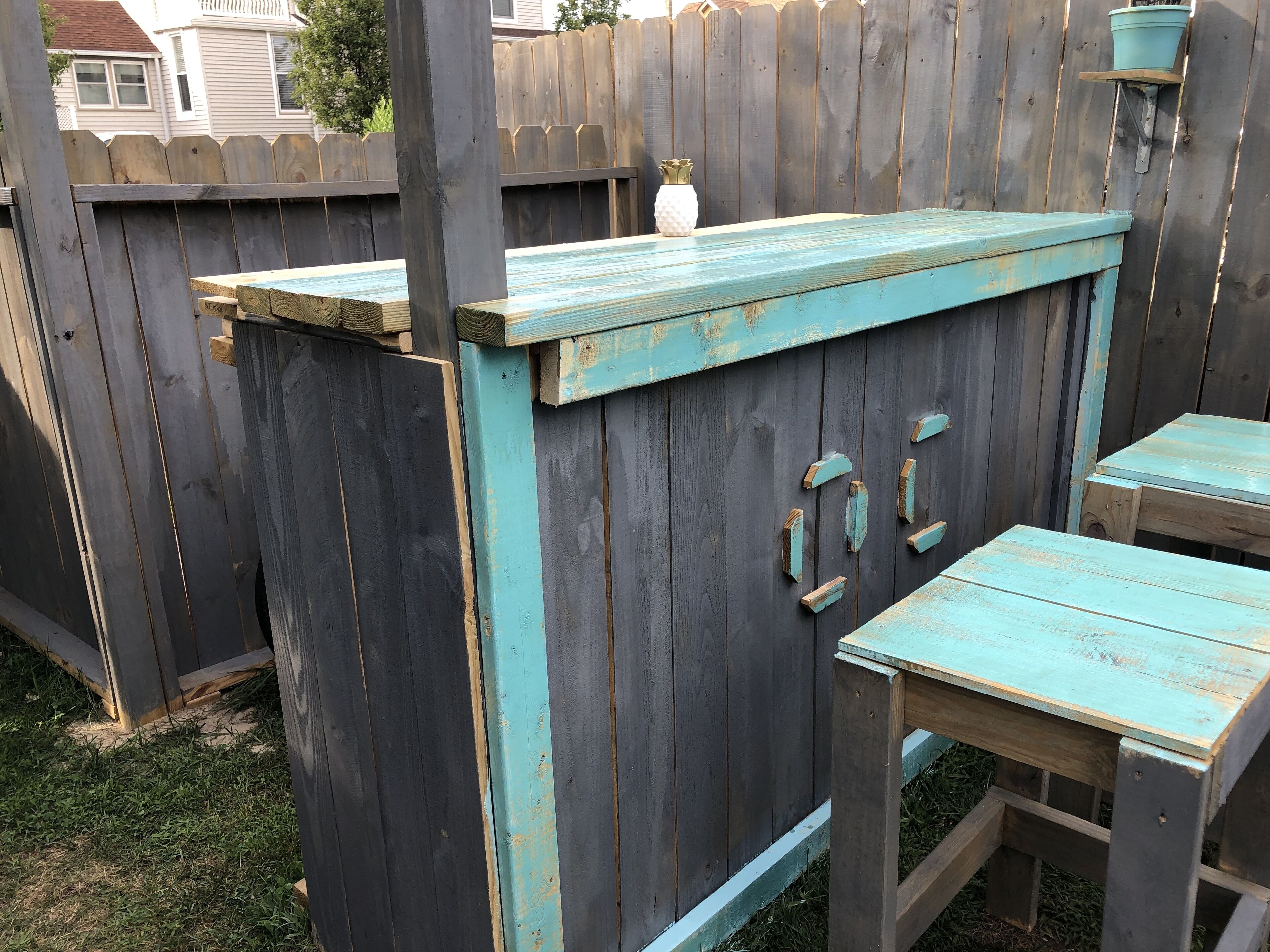 Un espace extérieur aménagé dans un style rustique, avec une table et des chaises en bois, un petit vase blanc et une clôture en bois en arrière-plan.