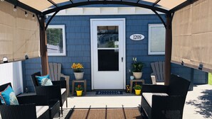 Un patio techado con mobiliario exterior, una puerta con un número y plantas en macetas.