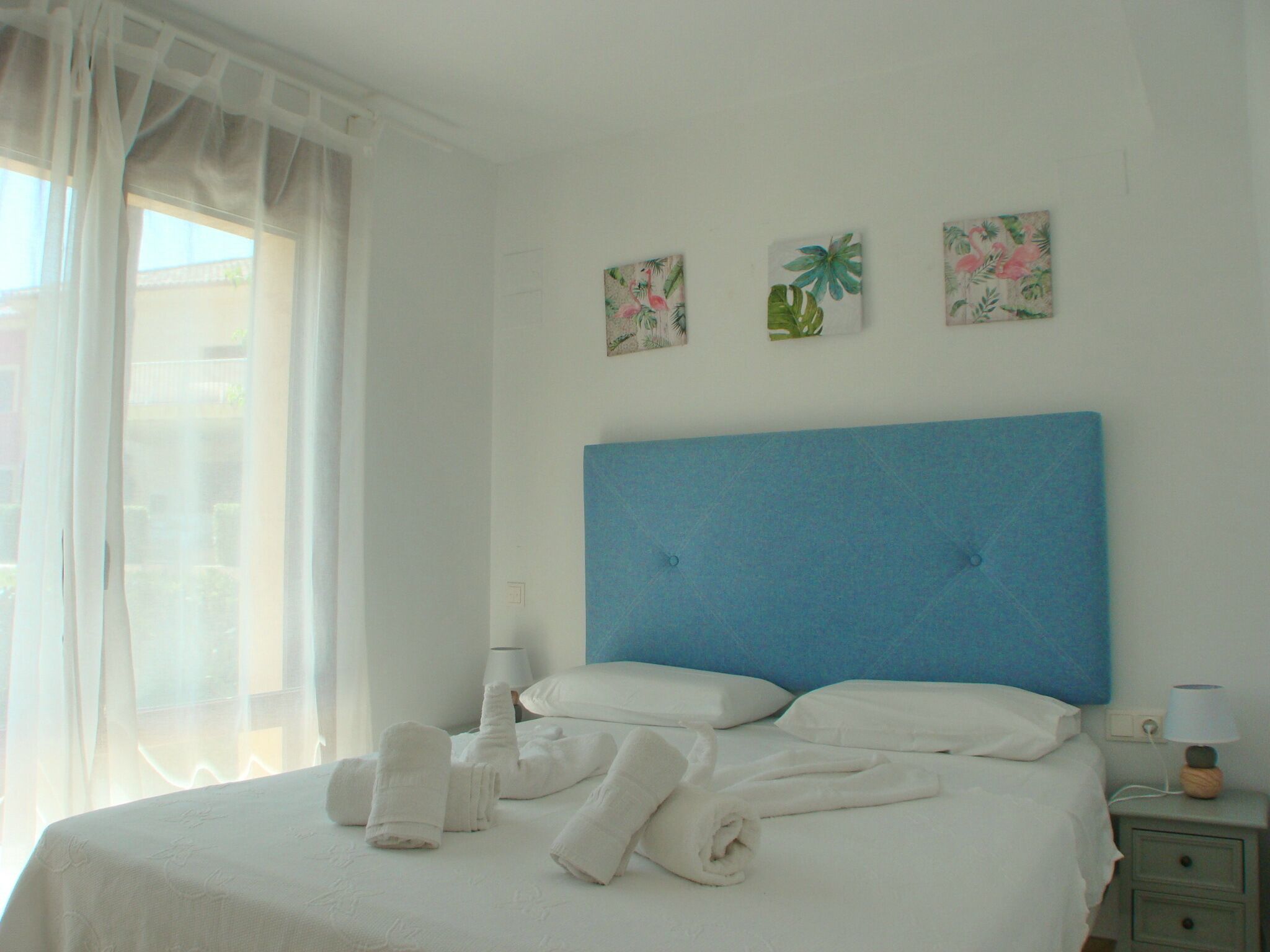 A hotel room with a large bed, white linens, a blue headboard, and three floral wall art pieces.