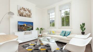 A modern living room with a glass coffee table, white sofas, a flat-screen TV, and a large window.