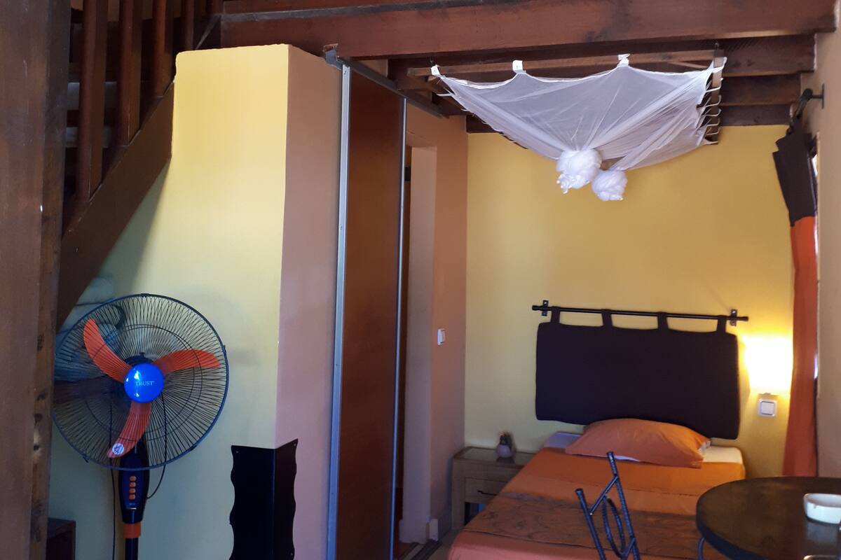 Un dormitorio con cama, mesita de noche, ventilador y mosquitera.