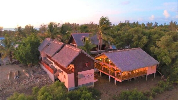 Una propiedad frente a la playa con varias edificaciones, incluyendo una casa principal y una construcción más pequeña con techo de paja.