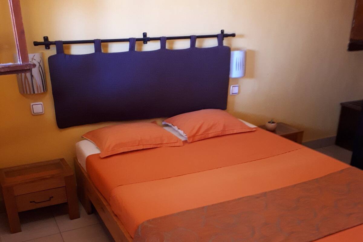 Una cama bien hecha con una colcha naranja y almohadas a juego, un cabecero de madera, una mesita de noche con una lámpara y un pequeño mueble con cajones.