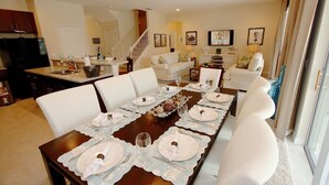 A dining room with a long table set for six, white chairs, and a kitchen in the background. - New Listing! Solterra Resort, 7mi to Disney & 17mi to Universal - IT2630 (Davenport)