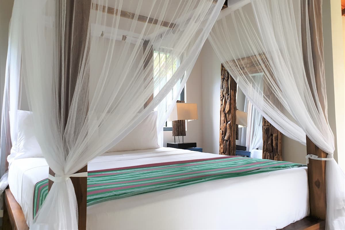Quarto com cama coberta por um dossel, mesa de cabeceira com abajur e cabeceira de madeira.