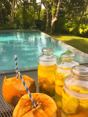 A poolside setting with a glass table holding a drink with a straw, a fruit, and two glass jars with a yellow beverage. - The Mugatiya (Ahangama)
