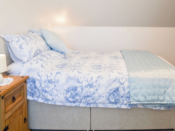 A bed with a floral patterned bedspread, a bedside table with a lamp, and a wooden nightstand with drawers.